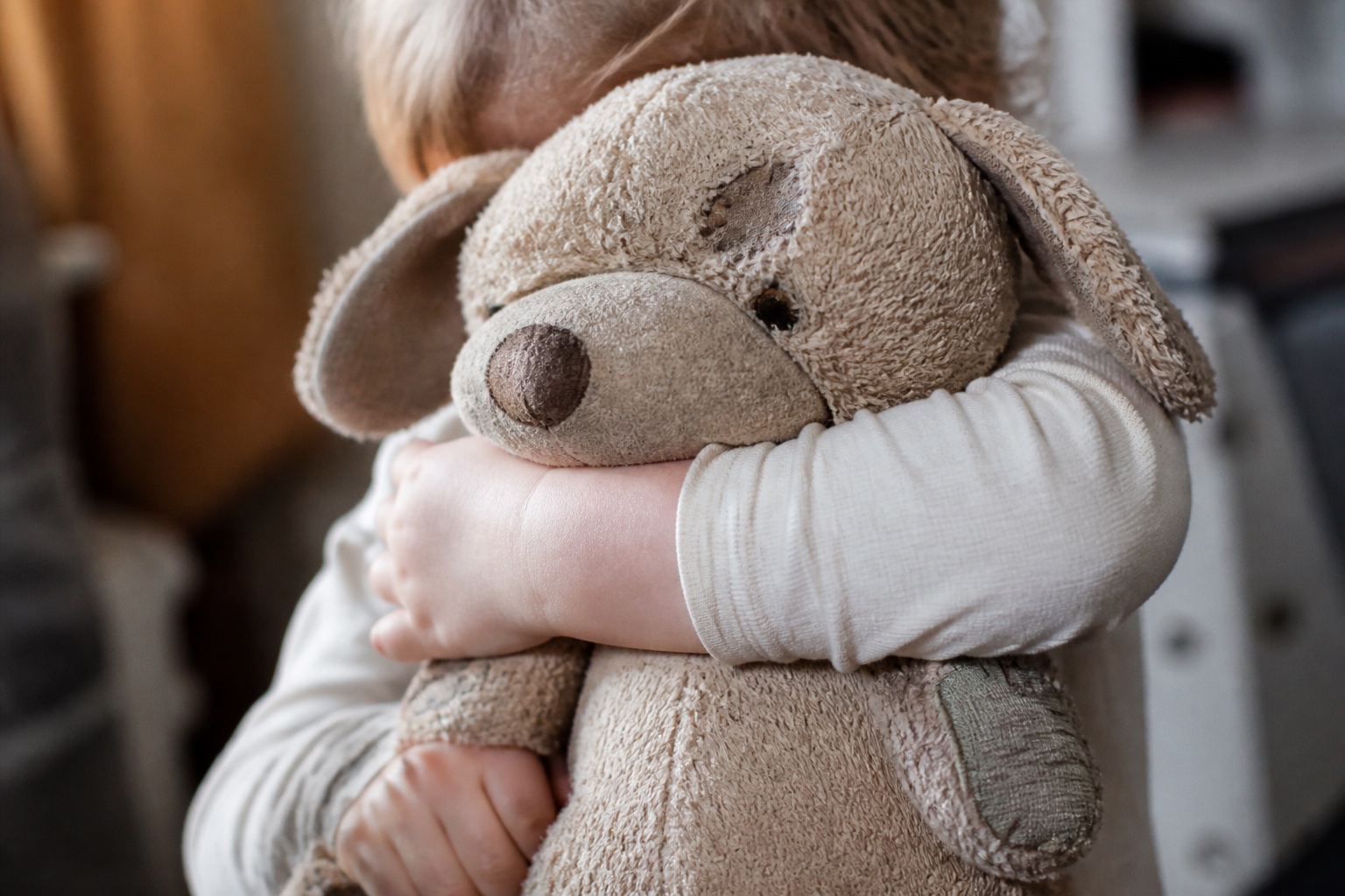 Ein brauner Teddybär sitzt auf einem zusammengerollten Schlafsack neben einem offenen Koffer mit gefalteter Kinderkleidung, Tageslicht von links, Nahaufnahme aus leichter Aufsicht.
